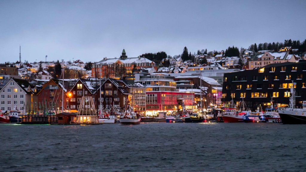Evening lights shimmer over the harbor of Tromsø, with wooden houses and fishing boats