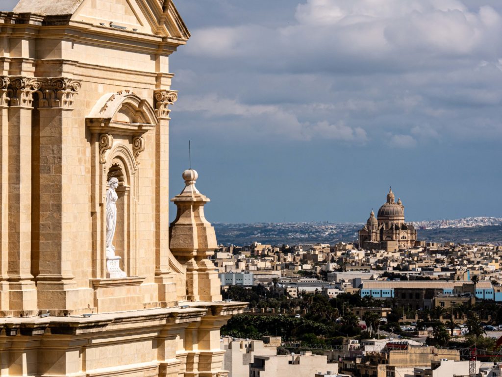 View over Gozo, Malta, sunlight brushing the limestone façade of a cathedral, with the Rotunda of Xewkija rising in the distance