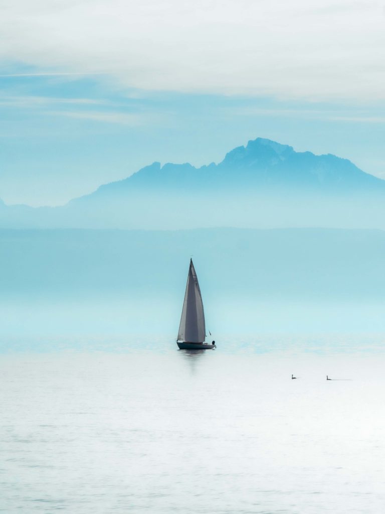 A lone sailboat drifts across the misty waters of Lake Geneva, beneath the distant Alps