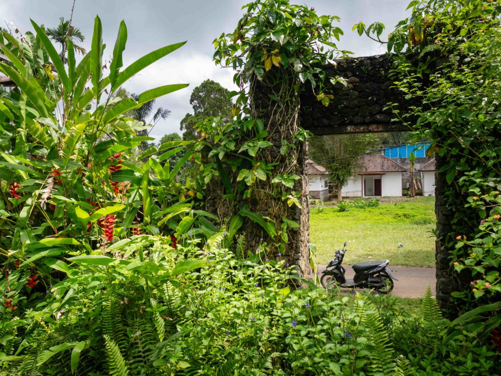 Stone gate covered with tropical plants overlooking a small village and parked scooter in central Bali