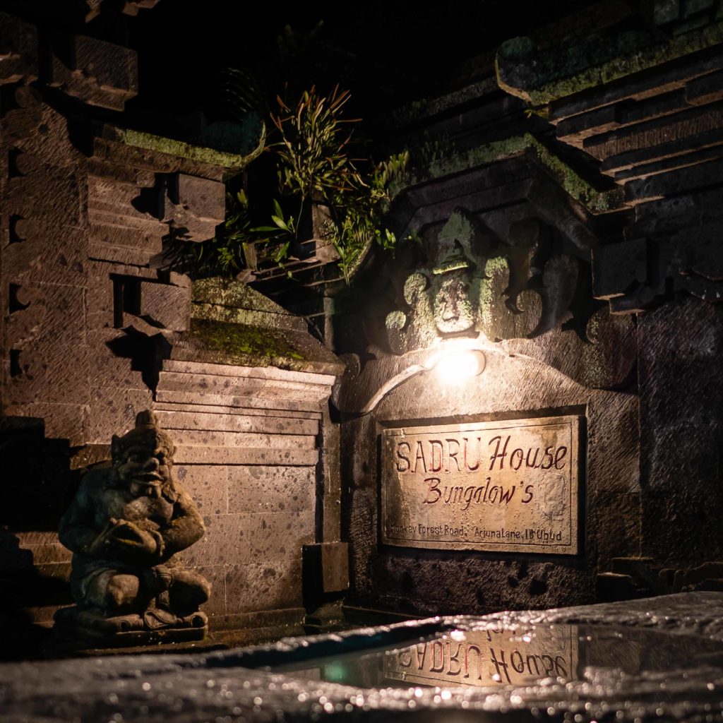 Night view of a traditional stone gate with guardian statue and sign for Sadru House Bungalows in Ubud